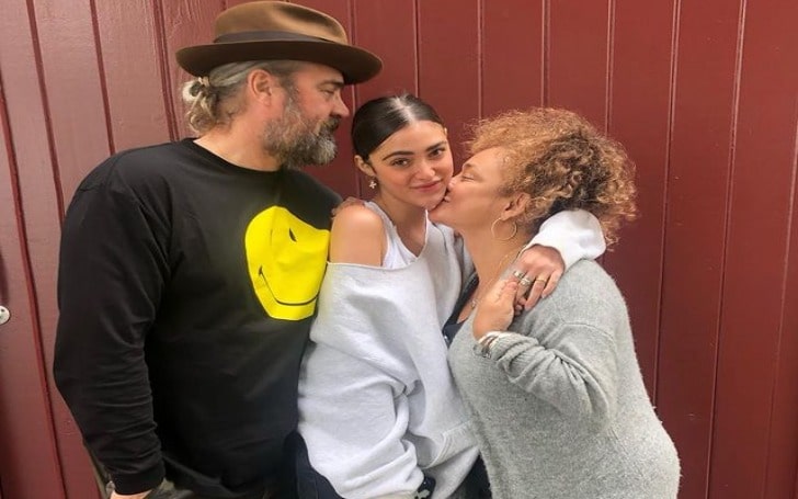 Luna Blaise in a white loose cloth with her father in a brown hat, black t-shirt with a yellow smiley and her mother in a grey cardigan.